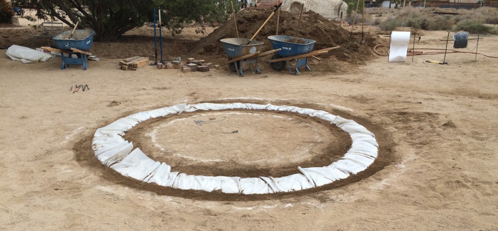 This is the foundation of a single dome that was used as part of a training video from Cal Earth. The required equipment (tampers, shovels, and wheelbarrows) are shown along with the rolls of plastic tubing and barbed wire used in construction and the pile of earth that will go into the tubes to form the walls.
